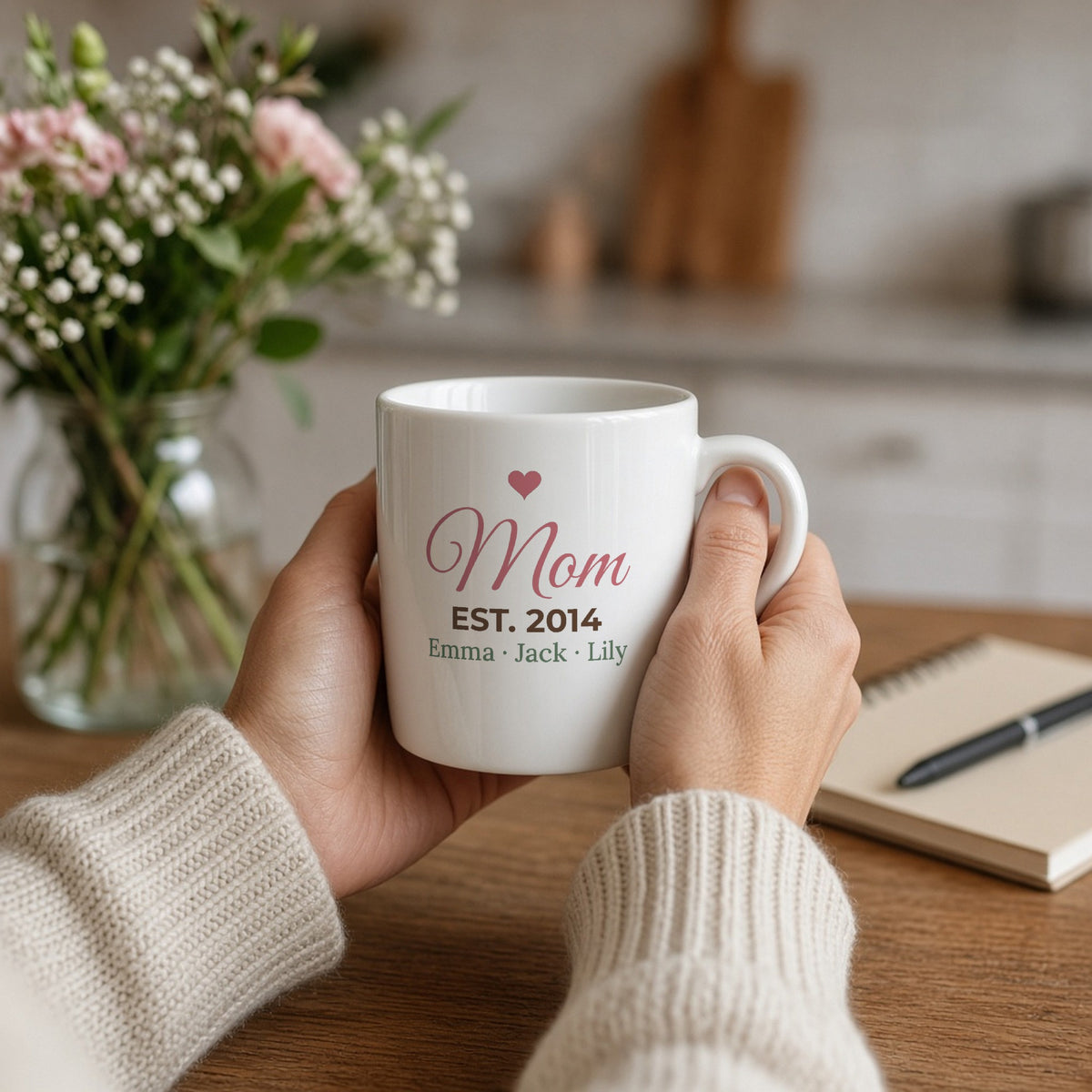 Personalized Mom Est. Year 11oz mug held in a cozy kitchen — lifestyle photo with the real print design. Shown with sample names.